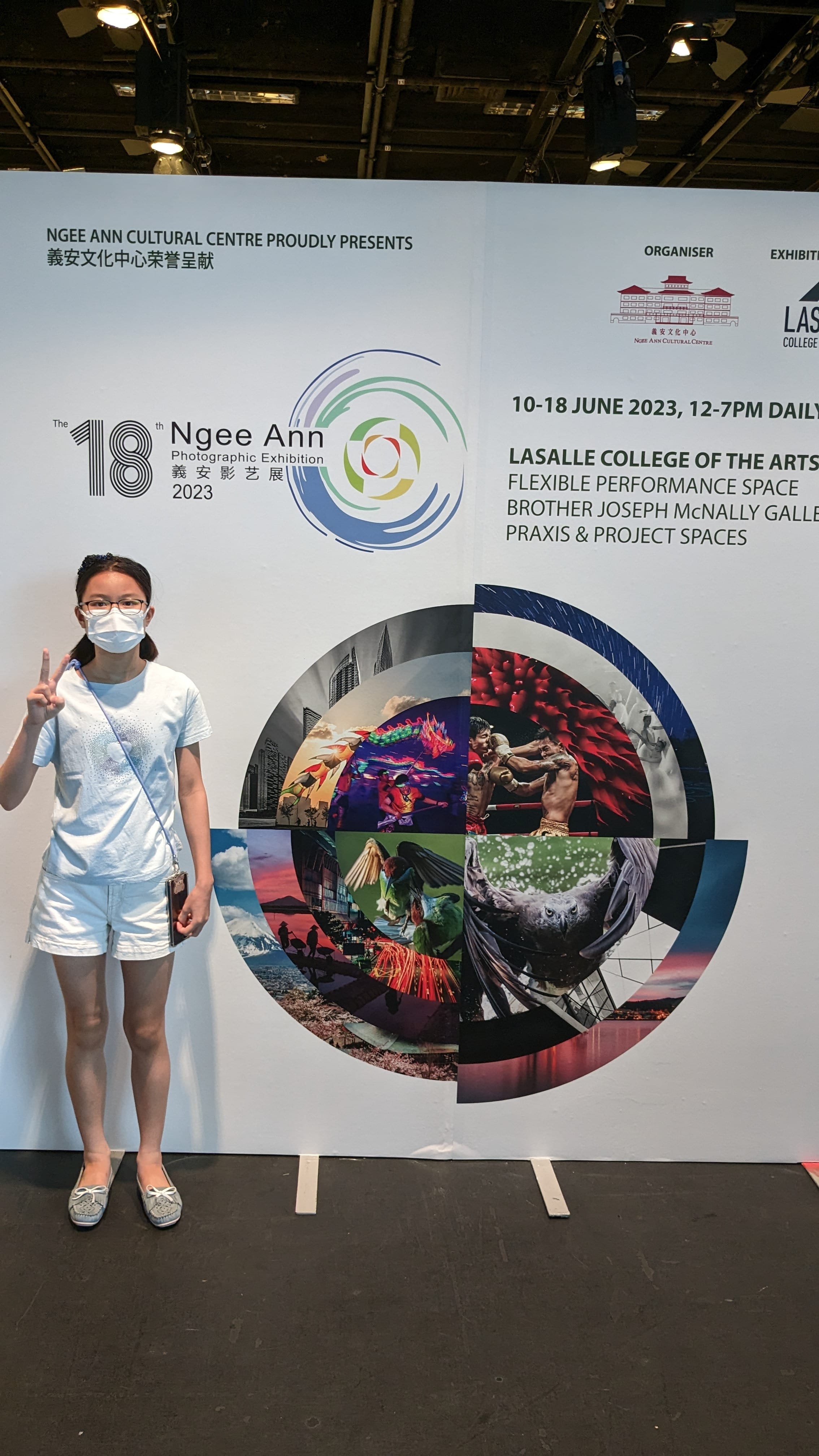 A masked young woman in white poses with a peace sign before the 18th Ngee Ann Photographic Exhibition 2023 banner.
